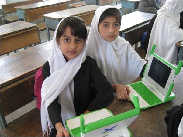 dos niñas afganas en su salon de clases muestran sus computadoras. son dos ceibalitas con las antenitas desplegadas, como las primeras que vinieron a uruguay. las niñas cubren parcialmente sus cabezas con telas blancas. usan camisas negras y mochilas. ambas miran a la camara y una sonrie levemente. tendran unos 9 años. 