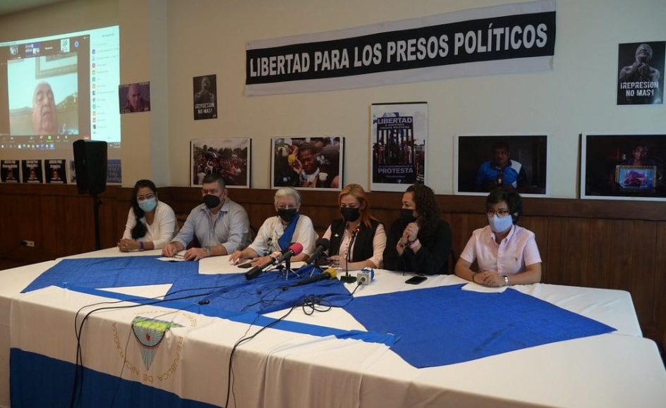 seis personas sentadas a una mesa para una conferencia de prensa, se ven los microfonos y la bandera nicaraguense sobre la mesa. en la pared un cartel dice libertad para los presos politicos.